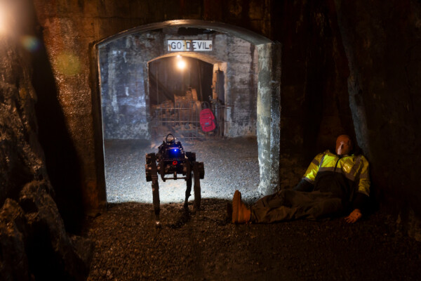 a legged robot walks down a mine past a dummy wearing a yellow vest and a red backpack that is hanging from a metal fence nearby