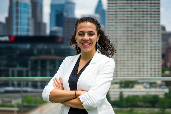 Director of Operations Sarah Parks of the men's basketball team staff folds her arms while posing with the Philly skyline in the background.