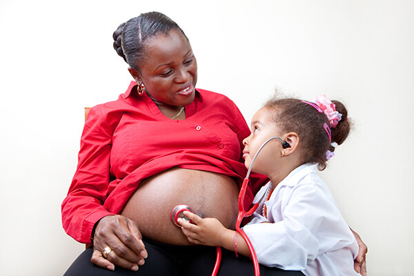 A young child wearing a doctor coat and stethoscope listens to the belly of a pregnant woman smiling down at the child.