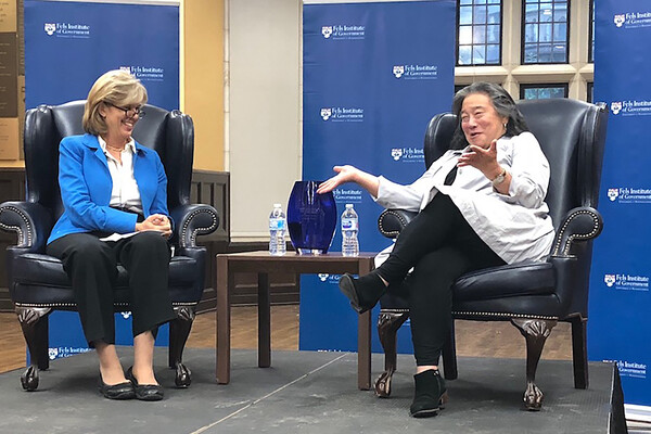Two people sit in wing-backed armchairs, separated by a small side table with an empty vase and two plastic water bottles, smiling and having a conversation in front of a wall with the words Fels Institute of Government and the Penn logo printed on it.