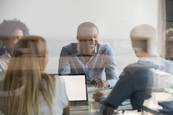 Four people sit at a table at work, one with a laptop, one smiling at a colleague