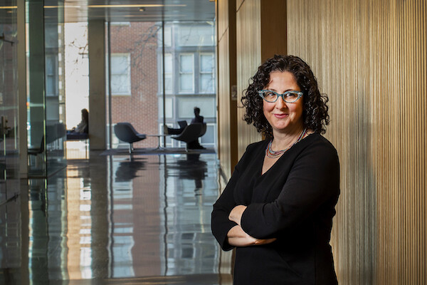 A person in a black dress and glasses stands against a wooden wall with arms crossed, looking at the camera, in the background is a huge window and people in chairs in front of the window.