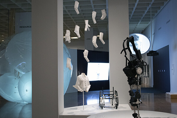 View of white garments hanging at the Designs for Different Futures exhibit, with a white shirt on a stand and a futuristic wheelchair and mechanical upright walking device on display.