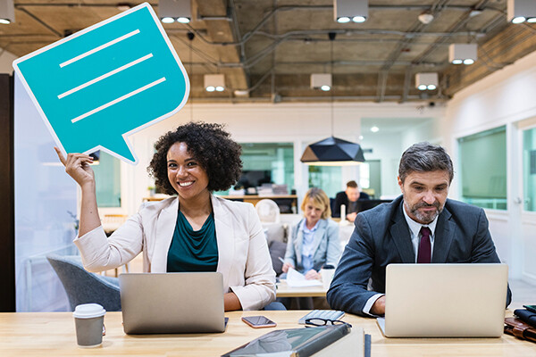 A woman and a man sitting side by side in an office working on laptops, she is holding an oversized "thought bubble" sign over her head and smiling