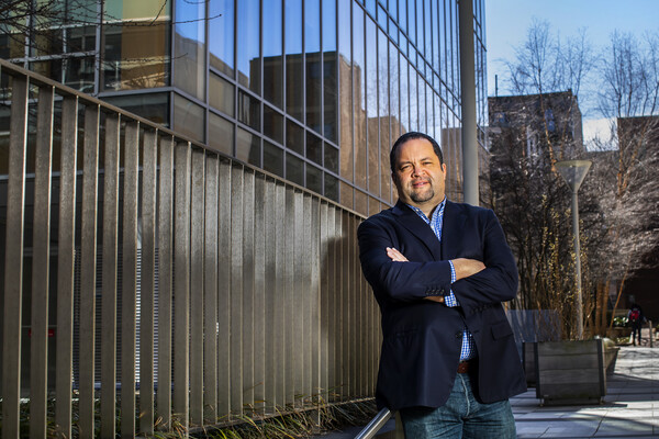 A person standing with arms crossed, in a courtyard next to a metal railing aside glass windows.