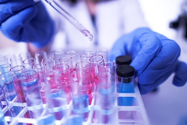 Gloved hands hold a dropper over a tray of glass vials in a lab