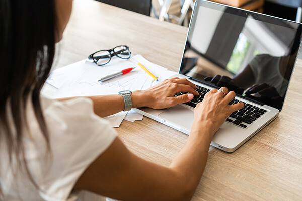 A person sits at a table typing on a laptop with papers and reading glasses and pens on the table