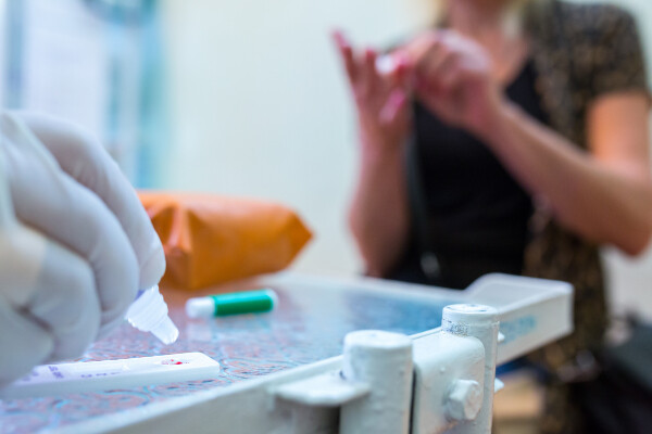 a gloved hand places a pipet tip over a plastic antibody test with a dot of blood, out of focus in the background a person puts a bandage on their finger