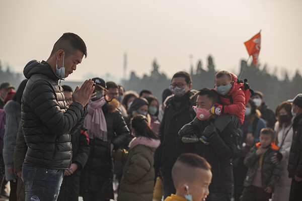 Person praying in front of a crowd in China with mask down around their chin, people wearing masks walk around them, outside a temple.