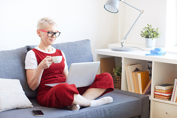Person sitting cross-legged on a couch holding a tea cup looking at a laptop