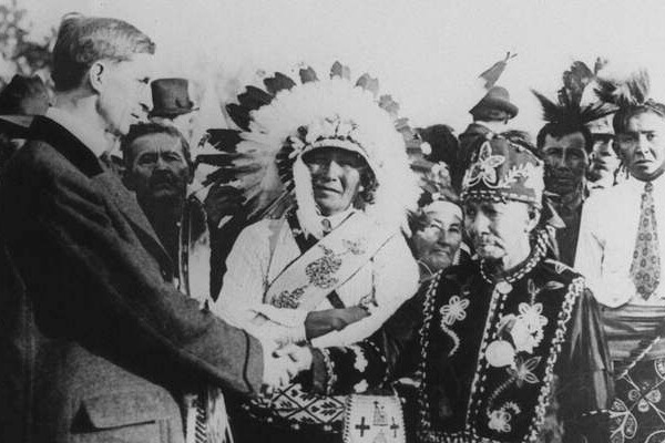 Man shakes hands with member of Ojibwe tribe of Wisconsin, an another man in headdress looks on.