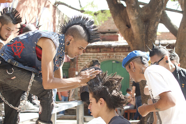 A young mohawked man with a leather vest featuring a red anarchy symbol styles another young man's hair into a mohawk