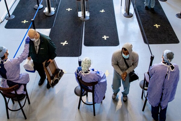 The entrance to a hospital. People in personal protective equipment swab others as they enter the building.