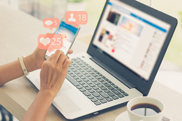 Hand holding a smartphone in front of an open laptop pressing oversized buttons for liking things on social media