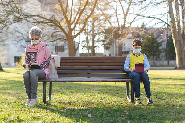 A grandparent and a child sit on a park bench in daylight, both wearing masks and sitting six feet apart.