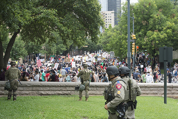 Armed soldiers stand in the grass in front of a low wall behind which a large protest is taking place.