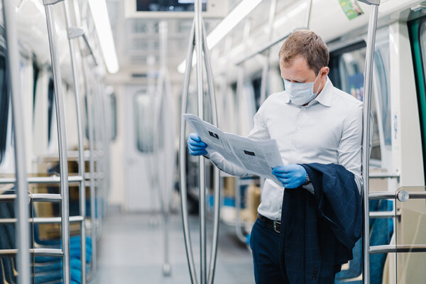 Person wearing face mask and latex gloves stands in an empty subway car holding a newspaper.
