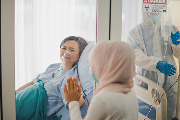 Person in hospital bed with COVID-19 presses their hand against a glass window with a loved one on the other side.