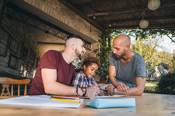 Happy same-sex  couple seated at a table outside homeschooling their young child.