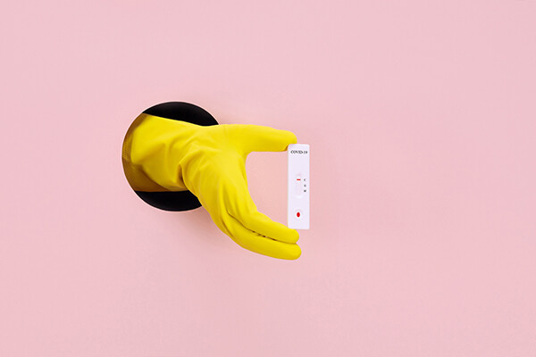 Rubber gloved-hand reaching through a hole in a wall holding a home COVID test.