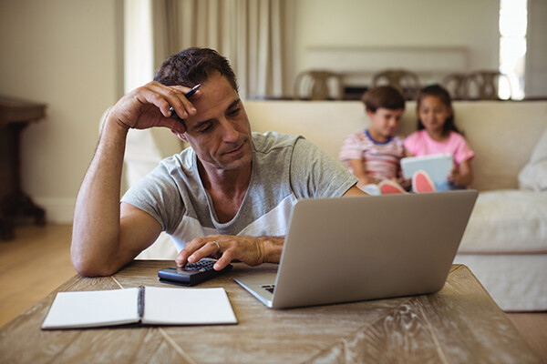 Parent at a coffee table looking at a laptop while holding a calculator, two small children sit on a couch behind them.