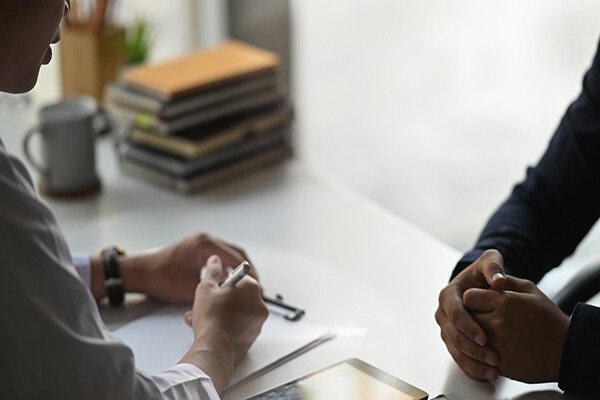 Closeup of a doctor and a patient at a desk, doctor is reviewing patient’s medical chart, patient has hands clasped.