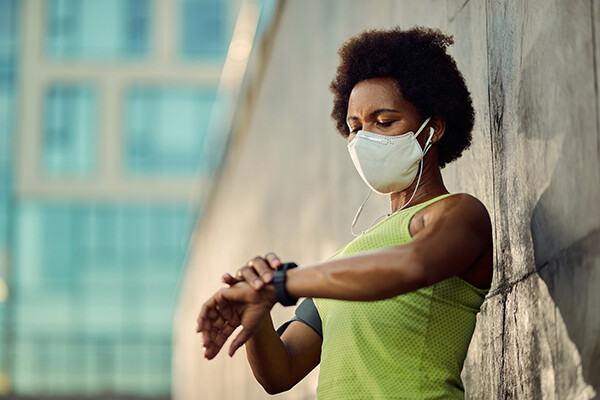 Person wearing a face mask leaning against wall and checking their fitness tracker on their wrist.