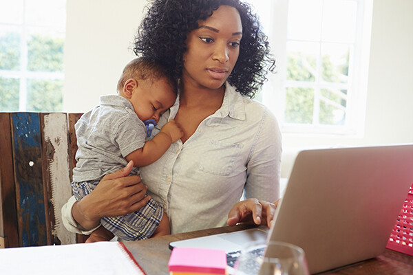 Parent holding a baby in one arm while sitting at a table with a laptop.