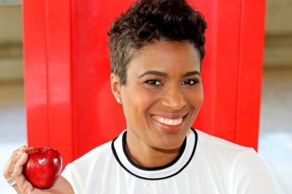 Smiling person holding an apple, standing in front of a red screen. 