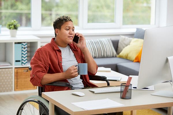 Person working from home, sitting at a desk with a computer, speaking on a smartphone, holding a mug.
