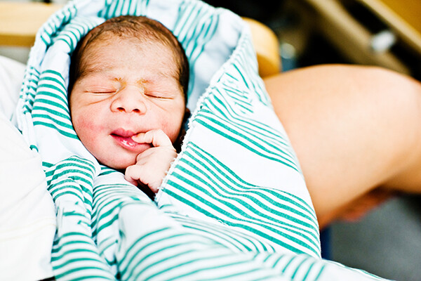 Newborn baby with finger in its mouth swaddled in a blanket.