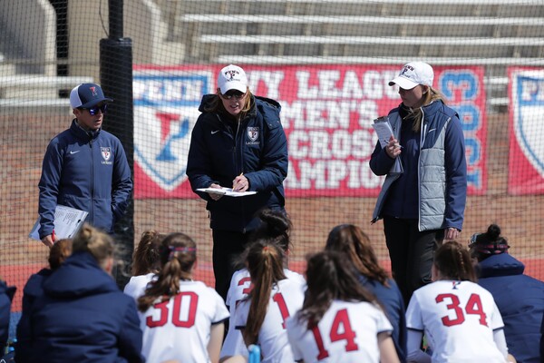 Standing between two coaches, Head Coach Karin Corbett addresses her players, who are seated at her feet.