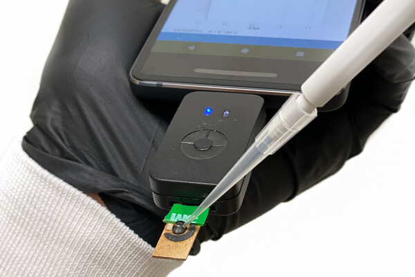 A hand in a black rubber gloves holding a cell phone with a sensor attached. A dropper is touching the sensor.