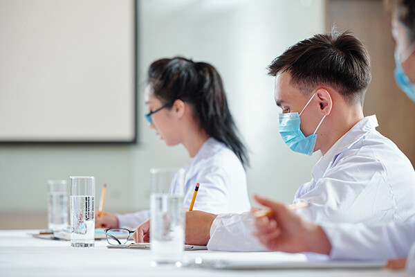 Medical students writing at a desk, one wearing a face mask.