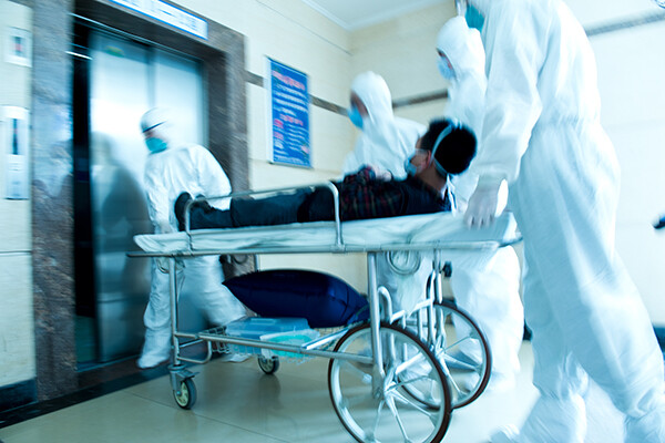 Three medical professionals in full PPE wheel a gurney with a patient towards elevator doors.