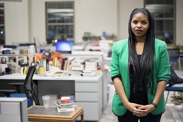 A woman with braids stands in an empty communal office space with books and computers on the desks