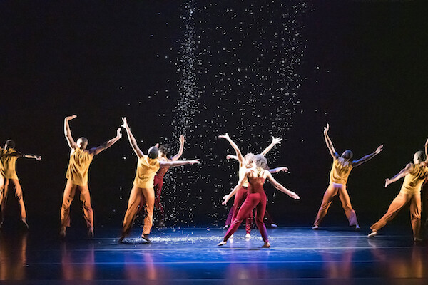Modern dancers with arms outstretched perform in a spotlight as confetti rains 