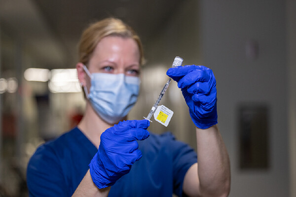 Medical worker wearing a face mask and latex gloves holds a syringe tagged with a vaccine label.