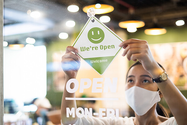 Person wearing a mask hanging a “We’re Open” sign on a window of a retail store.