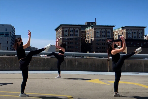 Three dancers wearing masks performing in an outdoor parking area.