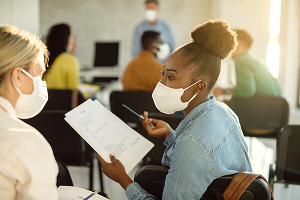 Two masked adult students in back of classroom discussing test results.
