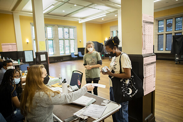 Two masked people standing and one poll worker seated at a table in Houston Hall.