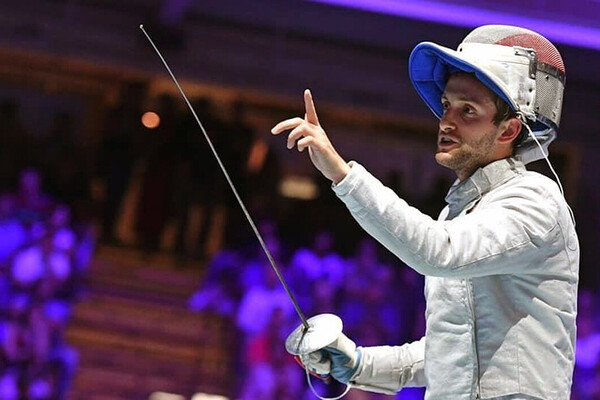 Shaul Gordon in fencing regalia holds his hand up while clutching a fencing sabre in the other.