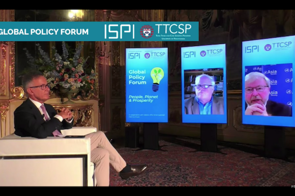 A man in a suit and tie and glasses sits in profile in a white chair as he looks at three video screens in front of him: one showing an illustration of a light buld and the words global policy forum, the middle screen showing a man with glasses in front of a book case and the last screen on the right showing another man with glasses, grey hair and a beard