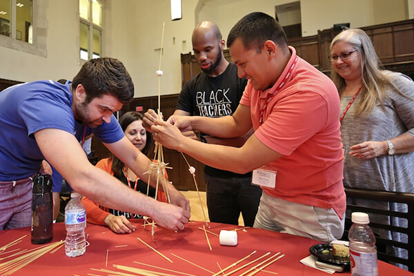 A group of educators in training work on a project building a tower of wooden sticks on a table.