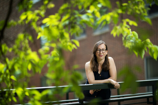 A person outside leaning over on a black railing, with leaves and trees blurry in the foreground.
