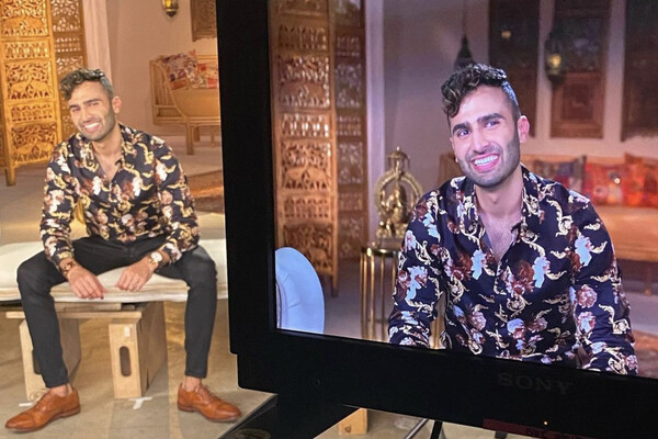 Dillon Patel seated and smiling behind a large television screen filming him.