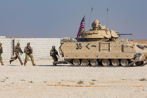 US soldiers holding guns are running behind a tank with an American flag on top in sand in Syria 