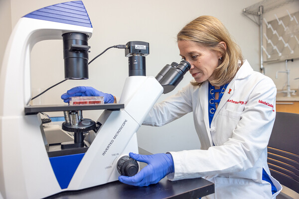 Sharlene M. Day looking in a microscope in a lab wearing a lab coat and latex gloves.