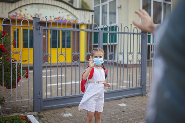 Kindergartner wearing a mask waves goodbye to parent in front of school gate.